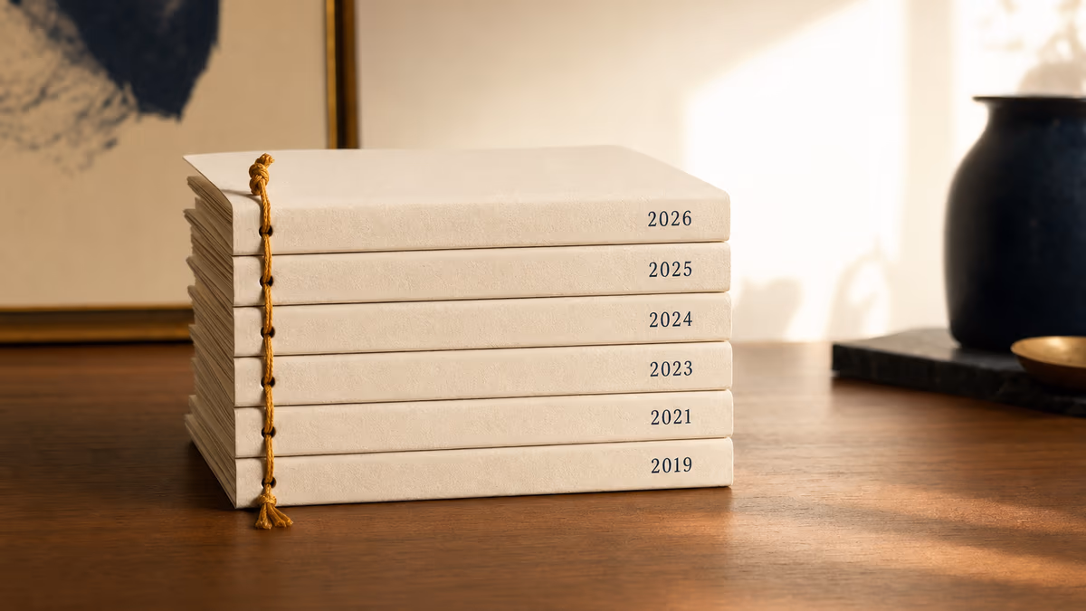 A precise vertical stack of six identical hand-bound cream booklets on a warm walnut desk, threaded by a single warm-amber waxed cord through each spine, with year stamps reading 2019, 2021, 2023, 2024, 2025, and 2026 — six layers of an ad stack made tactile.