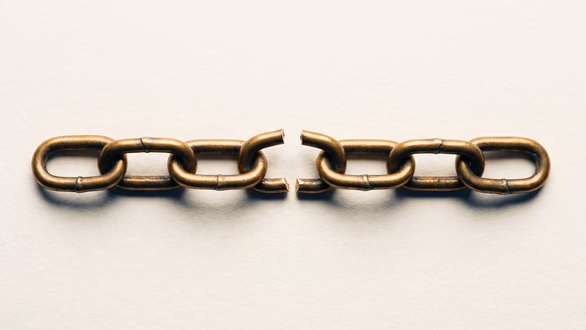 A short antique brass chain laid horizontally across a warm ivory cotton-fiber paper field, the central link cleanly broken into two halves with a small visible gap separating the two sides of the chain, brass patina and tarnish visible at the seams of each link.