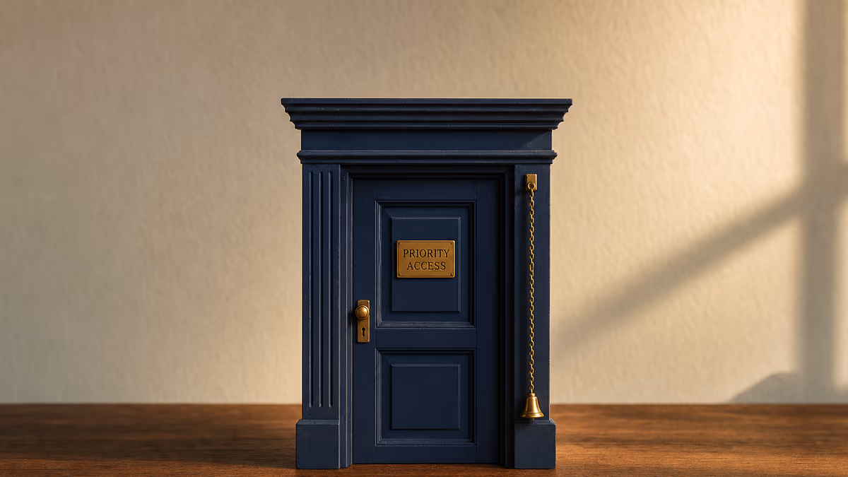 Editorial product photograph on a warm walnut surface showing a small handcrafted wooden doorway and door frame mounted upright, the door closed and painted matte deep editorial navy. On the door's central panel, an inset rectangular brass plaque is engraved in tabular small-caps warm amber reading PRIORITY ACCESS. To the right of the door, a single thin brass bell-pull chain hangs straight down from the upper frame, terminating in a small bell. Late-afternoon side-light from camera-right catches the brass plaque.